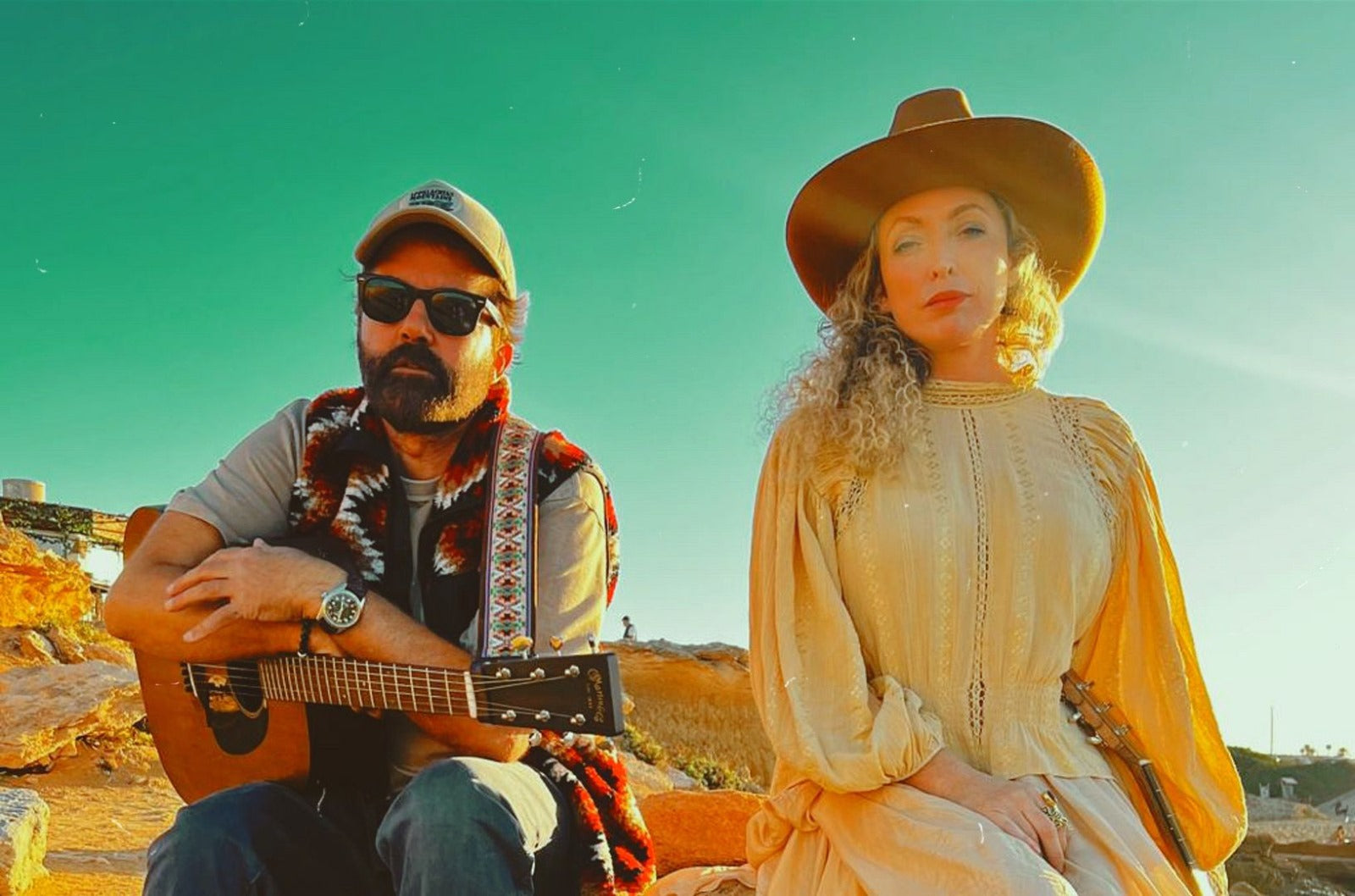 The band The Ting Tings, consisting of a man and a woman, are pictured outdoors against a clear blue sky to promote their new album, 'Home.' The man, wearing sunglasses, a cap, and a colorful vest, is holding an acoustic guitar, while the woman, in a wide-brimmed brown hat and a vintage beige dress, gazes confidently at the camera. The warm sunlight casts a glow on them, creating a relaxed and earthy atmosphere in the natural setting.